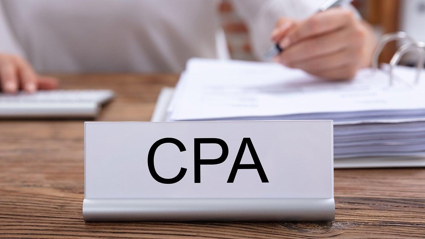 Close-up of a CPA nameplate on an office desk with a business professional reviewing financial documents in the background, representing year-round tax planning and financial guidance for small businesses in London, Ontario.
