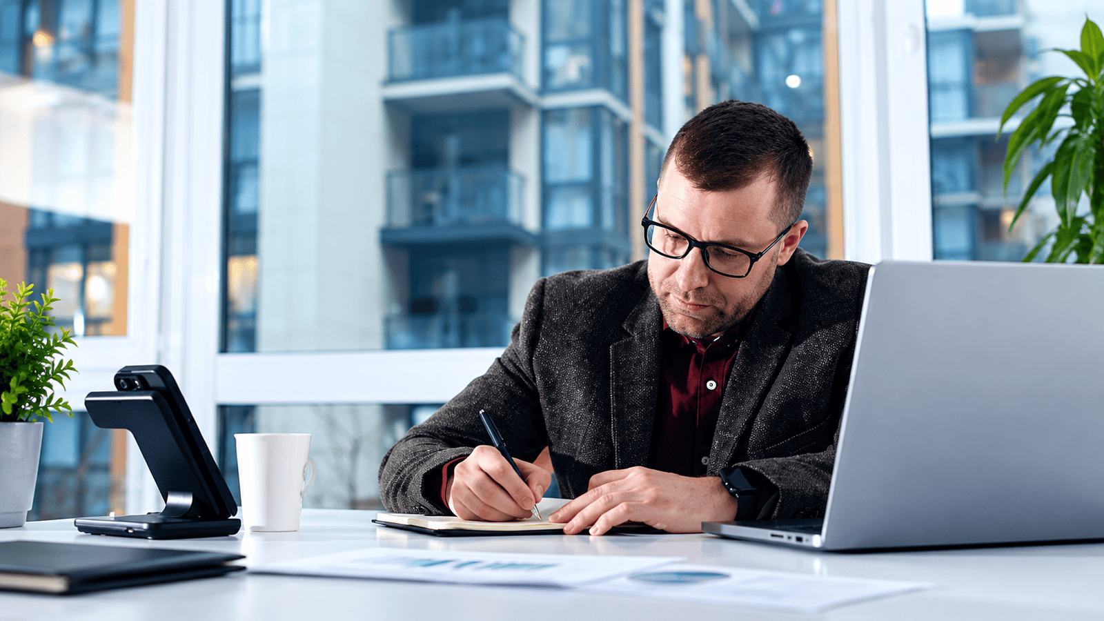 Business owner in glasses writing notes beside a laptop and financial reports in a bright office, representing the careful documentation behind family payroll for corporations across Ontario and Canada.
