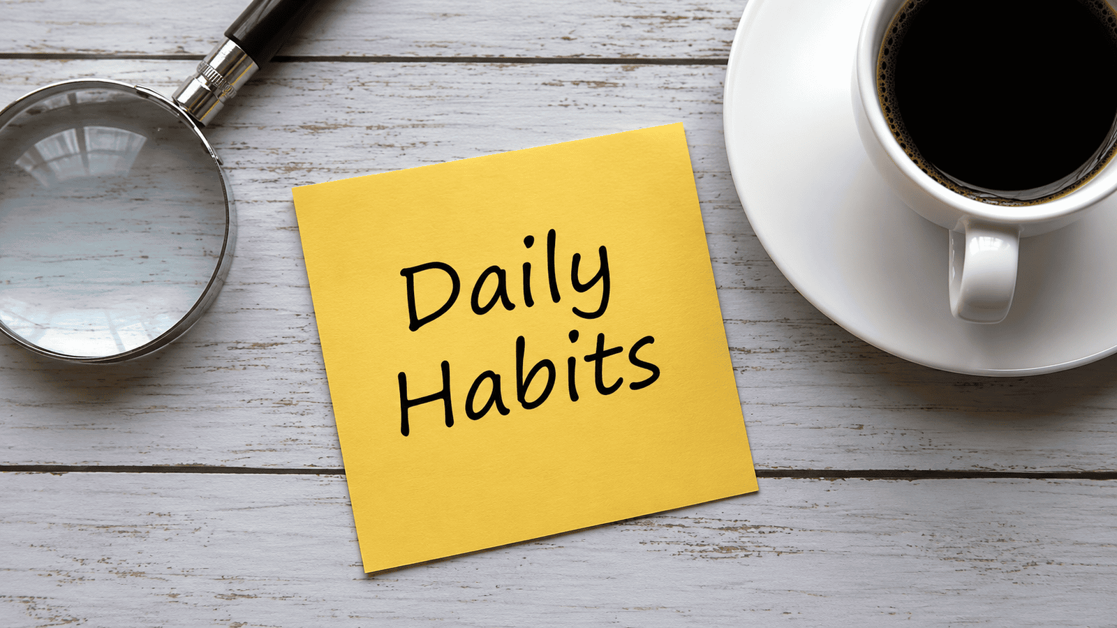 Overhead view of a yellow sticky note reading "Daily Habits" beside a magnifying glass and coffee cup, symbolizing the consistent bookkeeping and financial routines that help Canadian businesses stay audit-ready.