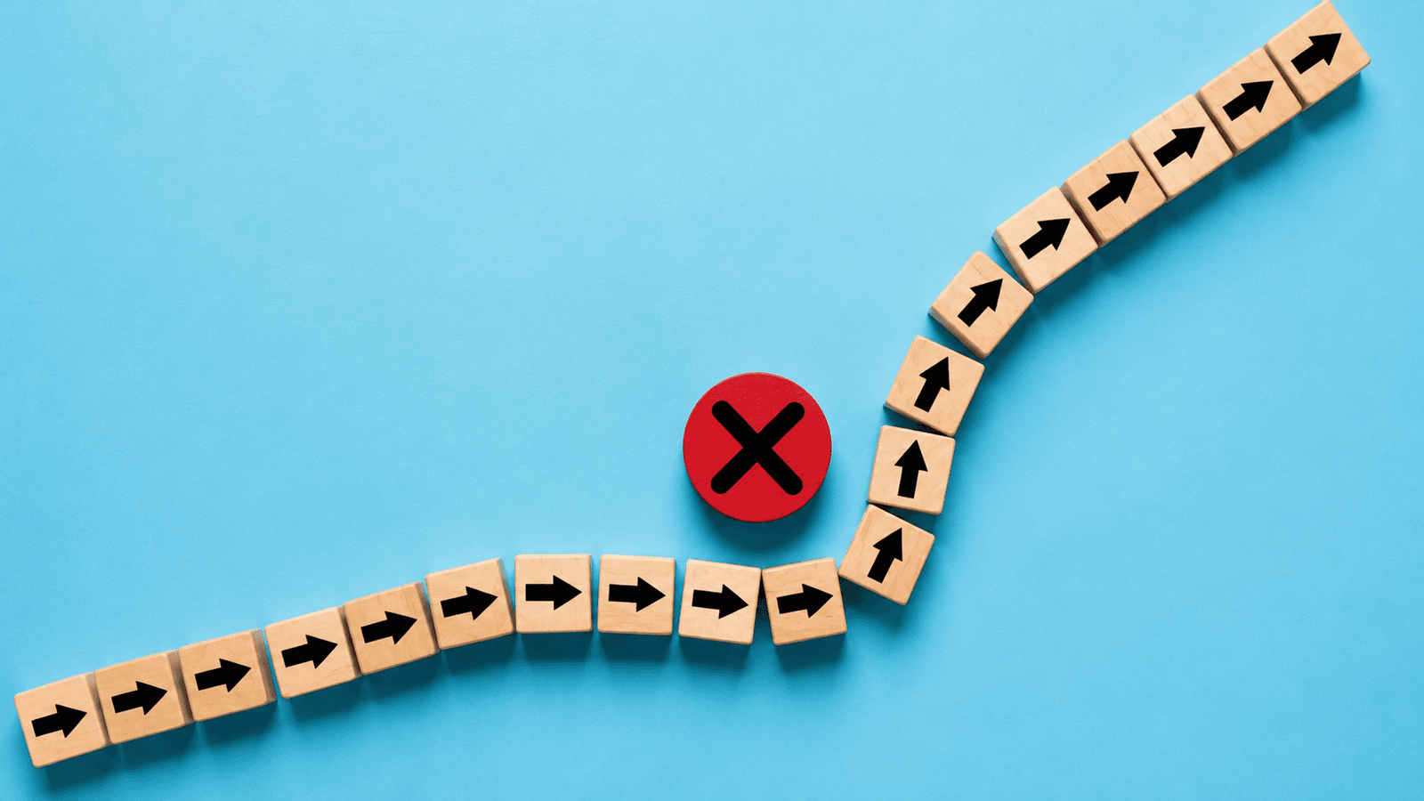 Minimal conceptual image of wooden arrow blocks changing direction around a red stop symbol on a blue background, representing common business mistakes, roadblocks, and the need to adjust processes for better outcomes.
