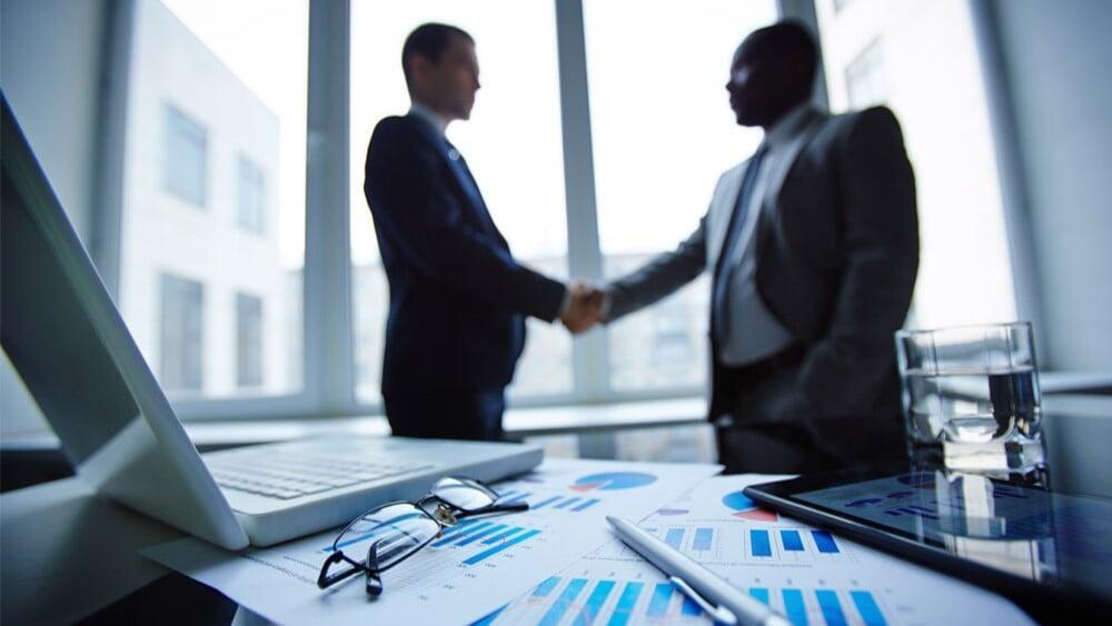 Close-up of two professionals shaking hands over a desk with financial charts, laptop, and documents, representing formalizing a business structure and making strategic decisions around incorporation and long-term growth.